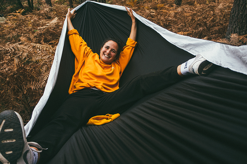 Eine Frau liegt lächelnd in einer schwarzen Campinghaengematte im herbstlichen Wald. Sie traegt einen gelben dicken Pulli, lange Hosen und Turnschuhe