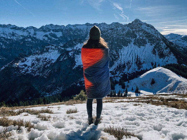 Frau in rot-blaue Campingdecke gewickelt steht auf einem Berg und betrachtet die Aussicht auf das Bergpanorama