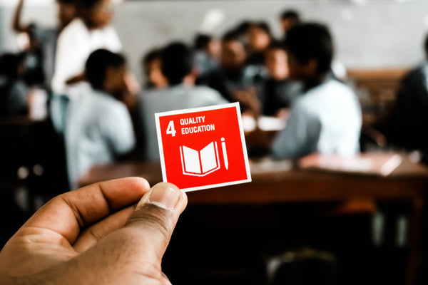 Hand streckt ein Kärtchen mit der SDG Nr. 4 aus. Im Hintergrund ist ein Klassenzimmer zu sehen. 