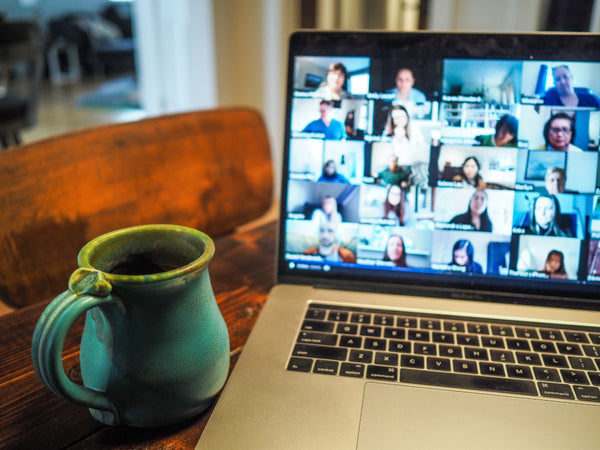 Laptop steht geöffnet auf dem Tisch, darauf zu sehen ist eine Online Video Konferenz. Daneben steht eine blaue Tasse.