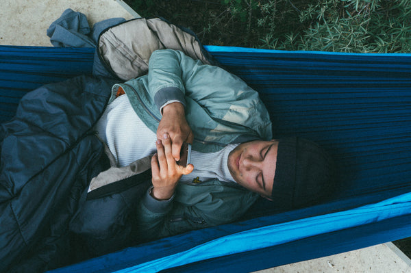 Mann liegt mit Handy und Schlafsack in blauer Hängematte