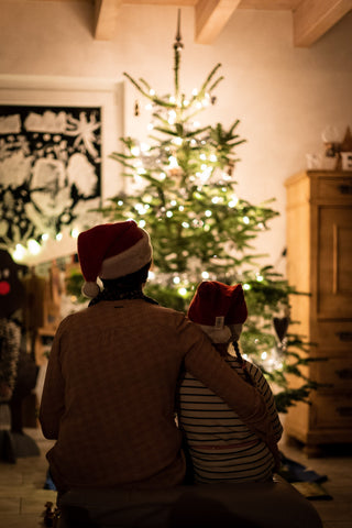 Mann und Kind Arm in Arm und Weihnachtsmütze auf dem Kopf sitzen vor dem geschmückten Tannenbaum