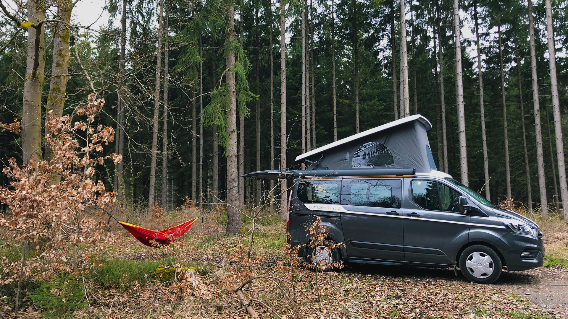 dunkler Campervan im Wald, daneben hängt eine rote Hängematte von HÄNG