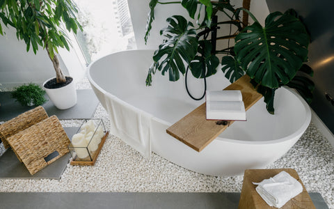A contemporary bathroom featuring a freestanding white bathtub surrounded by a pebble-covered floor. A lush green plant in a white pot adds a touch of nature, complementing the smaller potted plant on a gray shelf. Natural wood elements are seen in a bathtub caddy, a small stool, and a rectangular wooden tray holding candles. A wicker basket and a white towel on the wooden stool contribute to the spa-like ambiance. Large monstera leaves extend over the tub, enhancing the room's organic and tranquil feel.