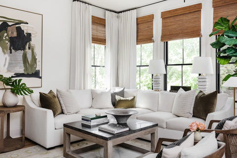 A cozy and stylish living room with large windows dressed in white curtains and topped with natural bamboo shades. A plush white sectional sofa is adorned with an assortment of decorative pillows in neutral tones. In front of the sofa is a modern coffee table holding books and a bowl, flanked by two contemporary table lamps with white shades on wooden side tables. A large abstract painting in earthy hues adds a touch of modern art to the room, and the space is complemented by indoor green plants, adding a vibrant pop of color. The room's décor is finished with a subtle patterned area rug and wooden accent furniture.