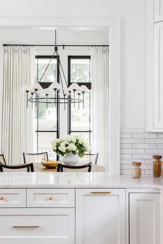 An elegant kitchen dining area illuminated by a modern chandelier with multiple white shades hanging over a round wooden table with black chairs. The table is beautifully set with a large vase of white hydrangeas at the center, complemented by a bowl of fresh lemons, adding a splash of color. Behind the dining set, large windows are framed by chic striped curtains, and the kitchen features white cabinetry with gold hardware, showcasing a clean and sophisticated design aesthetic. A white subway tile backsplash and a marble countertop add to the luxurious feel of the space.