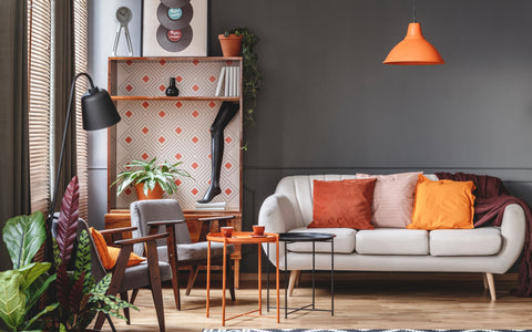 The image depicts a cozy living area with a modern aesthetic. There's a sleek white sofa adorned with orange and pink cushions, complemented by a dark maroon throw. An eclectic mix of furniture including a gray armchair, a set of three nesting coffee tables with orange and black frames, and a wooden dining set, contribute to the room's mid-century modern vibe. The space is accented with vibrant orange elements such as a pendant light and decorative pieces. A dark gray wall creates a contrasting backdrop, while natural light filters through wooden blinds, casting a warm glow. Houseplants add a touch of greenery, enhancing the room's inviting and stylish atmosphere.