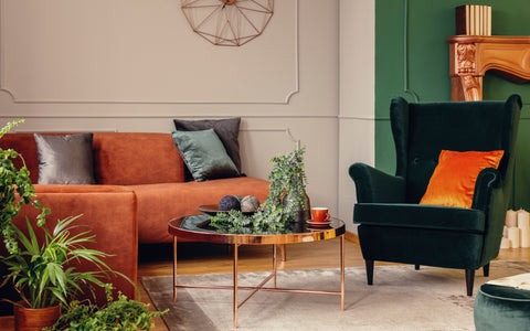 The image depicts a cozy living area with a modern aesthetic. There's a sleek white sofa adorned with orange and pink cushions, complemented by a dark maroon throw. An eclectic mix of furniture including a gray armchair, a set of three nesting coffee tables with orange and black frames, and a wooden dining set, contribute to the room's mid-century modern vibe. The space is accented with vibrant orange elements such as a pendant light and decorative pieces. A dark gray wall creates a contrasting backdrop, while natural light filters through wooden blinds, casting a warm glow. Houseplants add a touch of greenery, enhancing the room's inviting and stylish atmosphere.