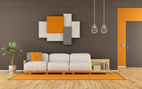 The image shows a minimalist living room with a contemporary design. The focal point is a clean-lined, white leather sofa with plush cushions, accented by a single orange throw pillow. Above the sofa hangs an abstract wall art piece featuring geometric blocks in shades of orange, white, and grey, complementing the bold orange accent wall to the right. The room is illuminated by two simple, exposed bulb pendants. A potted plant adds a natural touch to the space, and a small wooden side table with books suggests a comfortable reading nook. The flooring is a warm, honey-toned hardwood, with a large orange rug under the sofa, anchoring the seating area in this modern and stylish space.