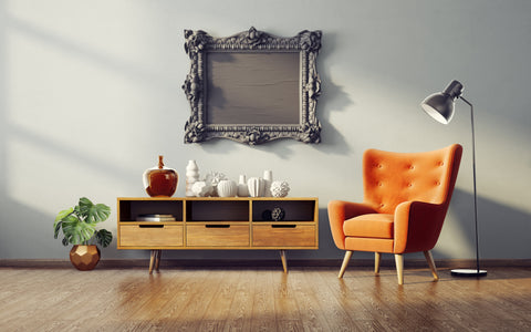 The image shows a stylish interior with a mid-century modern aesthetic. On the left, there is a vintage wooden sideboard featuring decorative vases in various geometric shapes and a large amber glass bottle. Above the sideboard hangs an ornate, baroque-style empty picture frame, adding a touch of classic elegance. To the right, a vibrant burnt orange armchair with tufted upholstery offers a cozy seating option, standing next to a minimalist floor lamp with a sleek design. A lush green plant in a geometric pot brings a refreshing burst of nature to the scene. The light hardwood floor and soft grey walls provide a neutral backdrop that highlights the warm orange tones and rich textures, creating a balanced and inviting space.