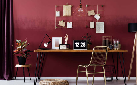 A contemporary home office space with a deep red wall. A sleek wooden desk is positioned against the wall, adorned with a digital clock showing 12:28, a small potted plant, and miscellaneous items including a hand model and framed pictures. Above the desk, a geometric wire grid holds notes and clippings. The workspace is complemented by a trendy gold metal chair and a round woven ottoman. To the right, a tripod floor lamp with a black shade stands beside a terrarium and a minimalist wire trash bin. The room's modern aesthetic is accentuated by a hanging copper geometric pendant light and coordinated decor items, creating a cohesive and stylish workspace.