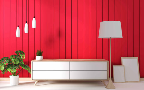 A vibrant and modern interior design with a bold red vertical slat wall. A Scandinavian-style sideboard with clean white drawers and a light wood finish sits against the wall, topped with a small potted plant on one end and a large white lamp with a fabric shade on the other. Above the sideboard, three sleek white pendant lights dangle at varying lengths, creating a dynamic visual line. Beside the sideboard, a lush green potted monstera plant adds a natural touch to the scene, while on the floor, two blank canvases lean against the wall, suggesting an artist's or decorator's work in progress. The room's bright color scheme and minimalist furniture present a striking contrast that's both eye-catching and stylish.