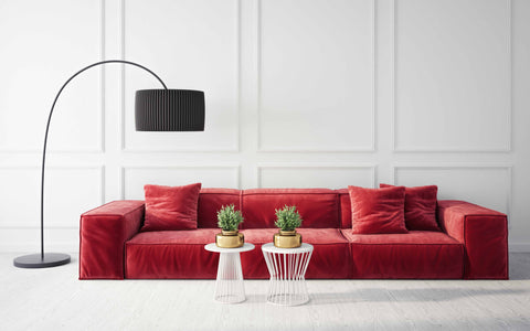 A minimalist living space featuring a striking red velvet sectional sofa with plush cushions, set against a classic white paneled wall. The sofa is accessorized with matching red pillows, enhancing its inviting appeal. In front of the sofa are two modern side tables, one white and one red, each holding a potted green plant atop a gold base, adding a touch of nature to the room. To the left, an elegant arc floor lamp with a sleek black base and a ribbed black lampshade creates a sophisticated lighting element that contrasts beautifully with the bright walls and sofa. The room's design is grounded on a light wooden floor that complements the clean, crisp aesthetic.