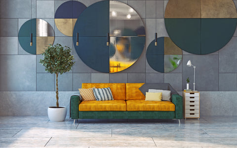 A contemporary living room featuring an eye-catching geometric wall decor with circular and semi-circular shapes in shades of blue, gold, and grey. A vibrant two-tone couch, with one half in a rich golden-yellow and the other in a deep teal, is adorned with decorative pillows. A potted indoor tree adds a touch of greenery, while a minimalist white floor lamp and a small side cabinet with drawers complete the scene, set against a textured grey wall and light wooden flooring.