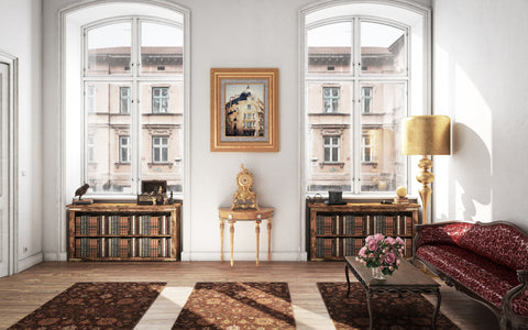 A sunlit vintage study with tall windows offering a view of historic buildings. The room is adorned with two bookcases filled with antique books, a small ornate gold-framed painting of a cityscape, and a classic wooden console table with gold accents. A luxurious gold floor lamp stands beside the window, casting a soft light across the room. The seating area features an opulent red and gold patterned chaise lounge, with a delicate glass coffee table holding a bouquet of pink roses in the center. Richly patterned rugs add warmth to the herringbone wood flooring.