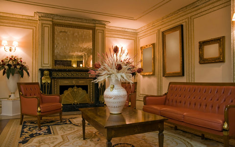 A classic and elegant living room interior with a sophisticated vintage charm. The room features a large, ornate mirror above a decorative black marble fireplace, flanked by golden sconces. Luxurious leather armchairs and a matching tufted leather sofa in a rich caramel color frame a dark wooden coffee table. A large vase with an arrangement of dried flowers serves as a centerpiece. The walls are adorned with gilt-framed mirrors, enhancing the room's grandeur, and the space is completed with an intricate cream and navy area rug on the floor.