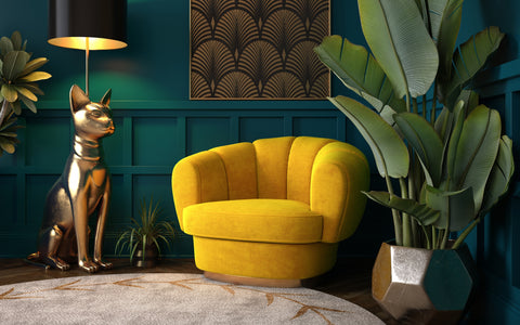 Alt text: "An elegant corner of a room featuring art deco style with a luxurious golden-yellow velvet armchair and a large metallic sculpture of a cat with a reflective surface. Behind the chair, a large tropical plant in a geometric planter adds vibrant greenery. The scene is set against a teal wall with paneled detailing, above which hangs a framed piece of art with a palm leaf pattern. A chic floor lamp with a black shade and gold interior casts a warm glow, highlighting the rich colors and textures in the room.