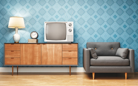 A stylish retro living room setup with a geometric blue wallpaper design. There is a vintage wooden sideboard with tapered legs, on top of which rests a classic white lamp, a pile of books, and an old-fashioned round clock. Beside it is an old-style television with dials and rabbit ear antennas. A modern touch is added by a sleek, dark gray mid-century modern couch with wooden legs and button details on the backrest.