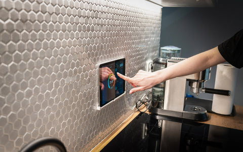 A person's arm reaching out to touch a built-in tablet control panel set into a hexagonal tiled backsplash in a modern kitchen. The tablet displays colorful smart home interface options including temperature settings. To the right of the tablet is a stainless steel espresso machine on a dark countertop, with a roll of paper towels and various kitchen utensils nearby. The kitchen's design is sleek with a combination of reflective metallic surfaces and warm wooden tones.