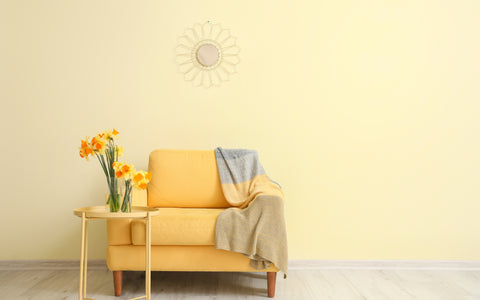 The image features a bright and cozy living space with a single-seater yellow couch, adorned with a textured gray and yellow throw blanket draped over one arm. A small round side table in a matching yellow shade holds a vase of fresh, blooming daffodils that complement the couch's vibrant color. Above the couch, on the pale yellow wall, hangs a decorative round mirror with sunburst metal frame, adding an artistic touch to the room. The scene exudes a cheerful ambiance, accentuated by the natural light that seems to be filtering into the room, creating a warm and inviting space.