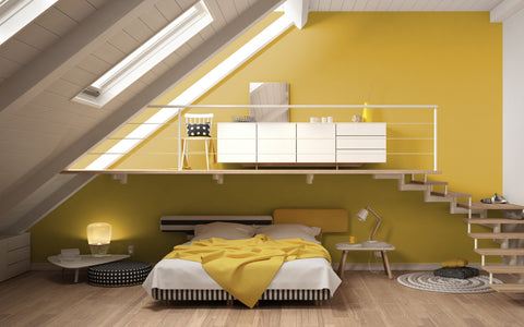 The image portrays a stylish, modern attic bedroom with vibrant yellow walls that create a focal point and add a burst of energy to the space. The room features a comfortable bed with a yellow headboard and a matching throw, contrasted by black and white striped bedding. On one side of the bed is a small round table with a contemporary lamp, and on the other side, a minimalist wooden chair and desk lamp. A black and white pouf, along with a round patterned rug, adds a touch of graphic design to the room. The upper level, accessible by wooden stairs, showcases a sleek white storage unit and a workspace with a chair. Skylights let in ample natural light, enhancing the warmth and welcoming atmosphere of the room.