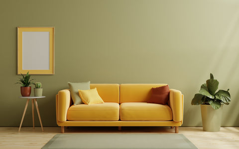 The image captures a tranquil living space defined by a contemporary aesthetic, featuring a sleek mustard yellow sofa adorned with cushions in shades of yellow and a rich terracotta. The furniture is set against a muted olive-green wall, which harmonizes with the sofa's warm tones. A minimalist gray side table displays a small collection of houseplants, including a cactus and a larger potted plant, introducing a refreshing natural element to the composition. An empty yellow-framed picture hangs on the wall, waiting to be filled with art or photography, and a subtle area rug lies beneath the sofa, grounding the setting. The room's overall ambiance is serene and stylish, with a thoughtful use of color and decor to create a cohesive and inviting space.
