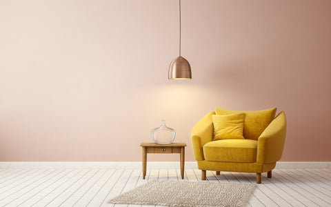 The image depicts a minimalist yet elegant interior scene featuring a luxurious mustard yellow armchair with plush upholstery and two matching pillows. The chair sits against a soft peach-pink wall, complemented by a white wooden floor. To the left of the armchair is a simple wooden side table holding an oversized clear glass decorative bottle. A modern pendant light with a copper finish hangs from the ceiling, casting a warm glow that enhances the room's inviting atmosphere. A shaggy off-white rug lies partially beneath the armchair, adding a cozy texture to the space. The overall composition of the room evokes a sense of contemporary sophistication with a pop of vibrant color.