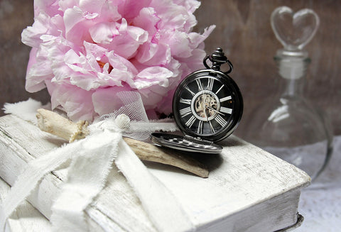 Im Hintergrund ein Brautstrauß in Rosa, eine Herz Vase und im Vordergrund ein altes Buch und eine alte Uhr. 