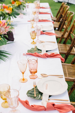 Ein langer, festlich gedeckter Tisch im Garten mit braunen Stühlen und korall farbenen Akzenten in der Tischdeko.