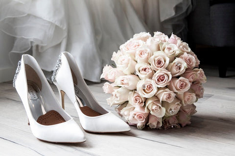 Eine Nahaufnahme von weißen Brautschuhen auf der linken Seite und einem Brautstrauß auf der rechten Seite. Im Hintergrund sieht man den Tüll des Brautkleides hängen. 