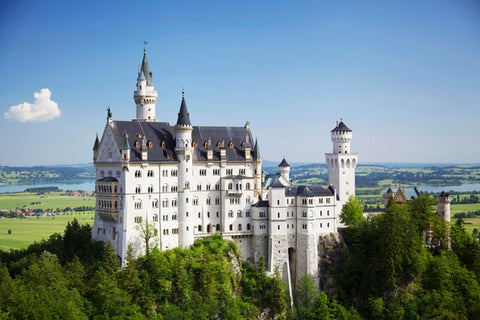 Ein prunkvolles, weißes Schloss mit Türmen ist umringt von Wäldern, Feldern und strahlend blauem Himmel.