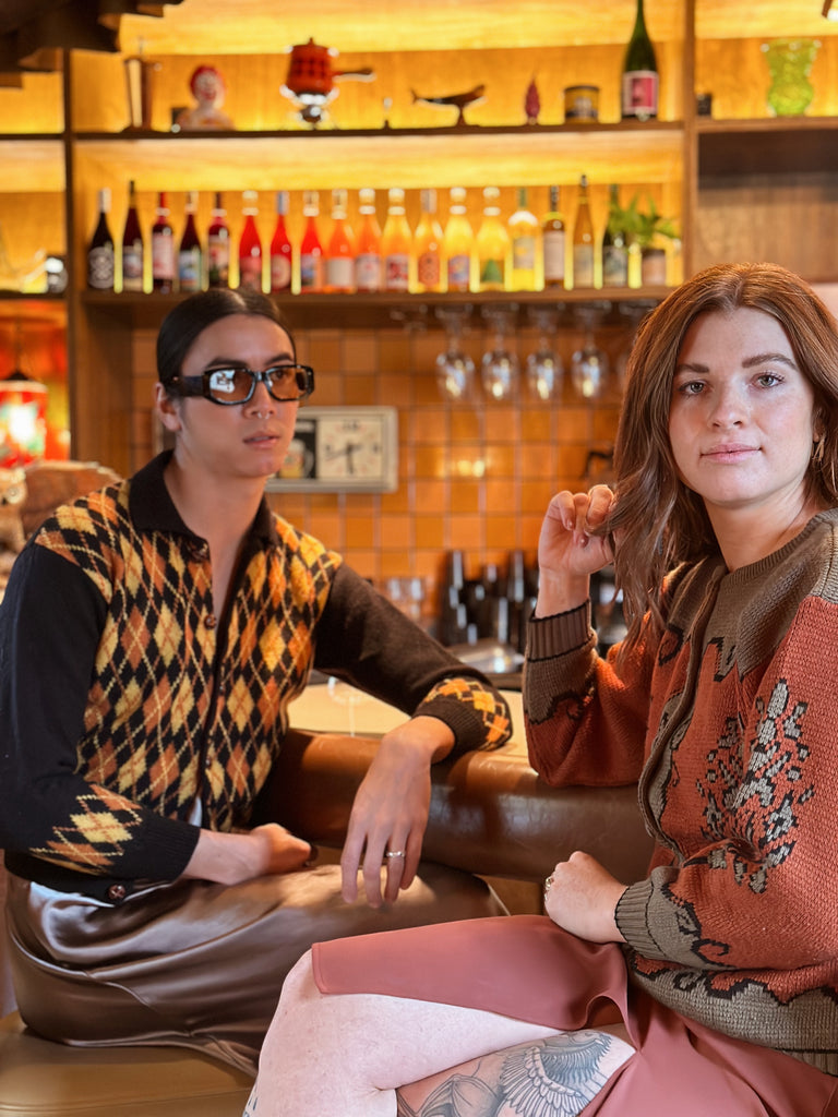 Two people sit at the bar of Class Clown wearing vintage 60s style cardigan sweaters