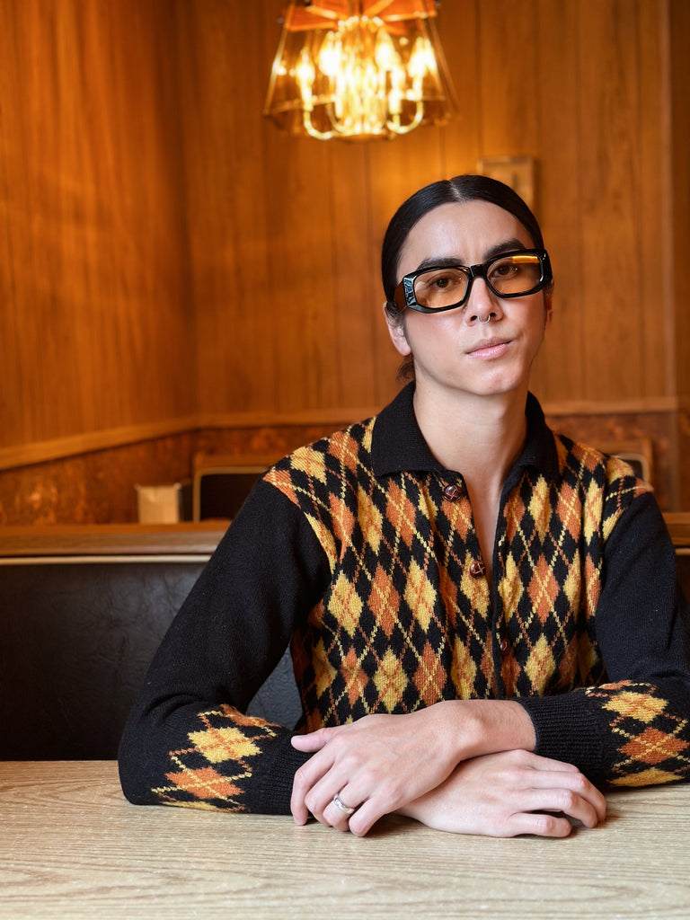 Person sits in a booth wearing a vintage orange, yellow, black argyle cardigan