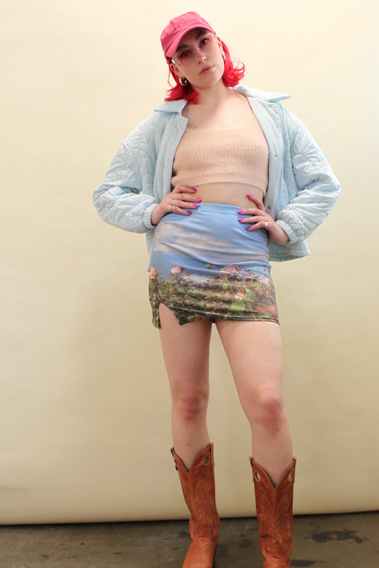 Woman wears a blue cloud jacket, pink crop tank, and graphic field print mini skit with brown cowboy boots