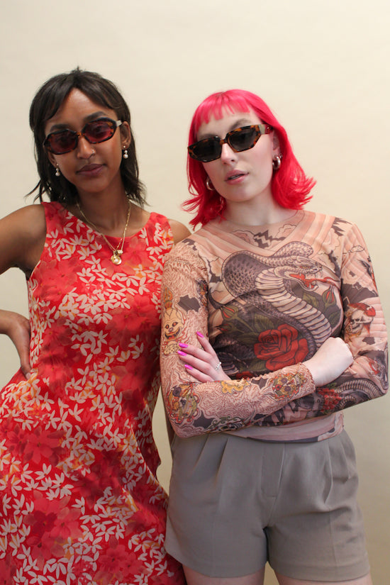 Two women wearing sunglasses one in a vintage floral red silk dress and one with pink hair in a graphic mesh shirt and shorts