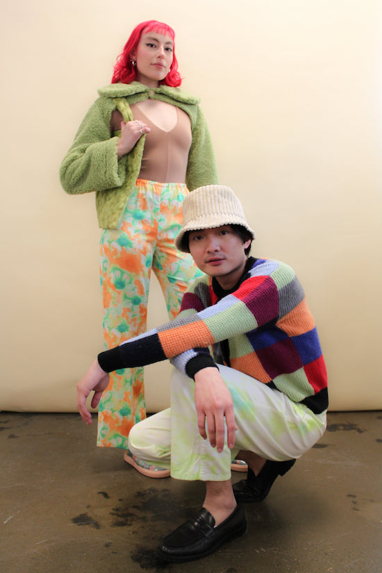 Person squats wearing a patchwork cardigan and tie dye pants in front of a woman wearing a green shrug jacket