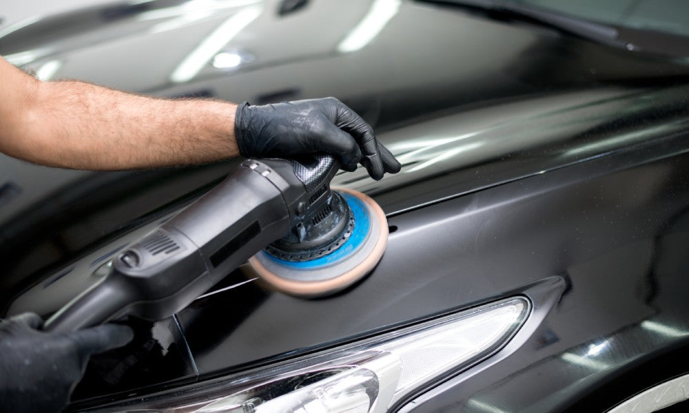 An auto technician wearing black protective gloves in an auto restoration shop polishing a black vehicle.