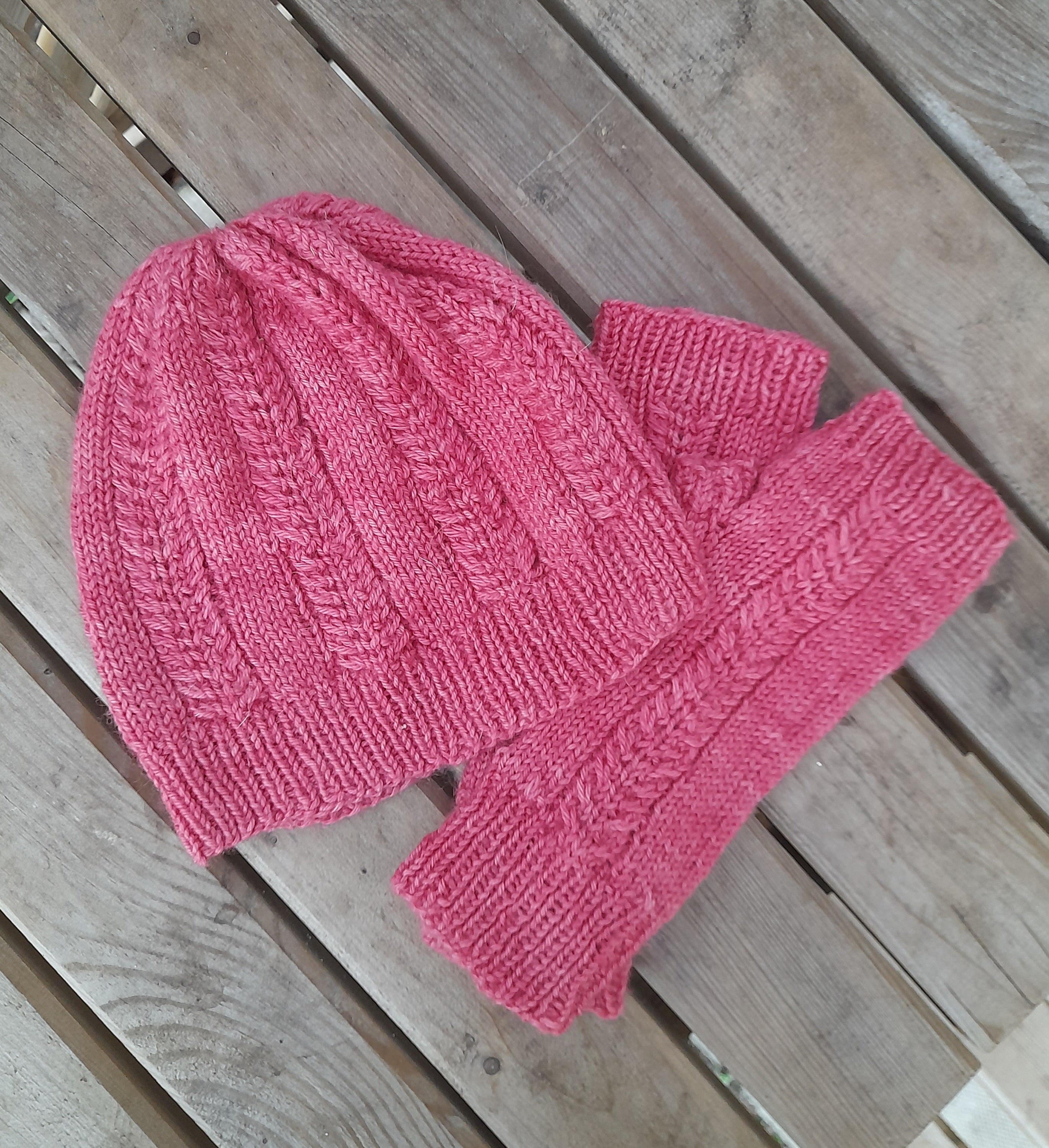 Pink knitted hat and fingerless mitts resting on a wooden bench. Both items feature textured stripe details running vertically along them