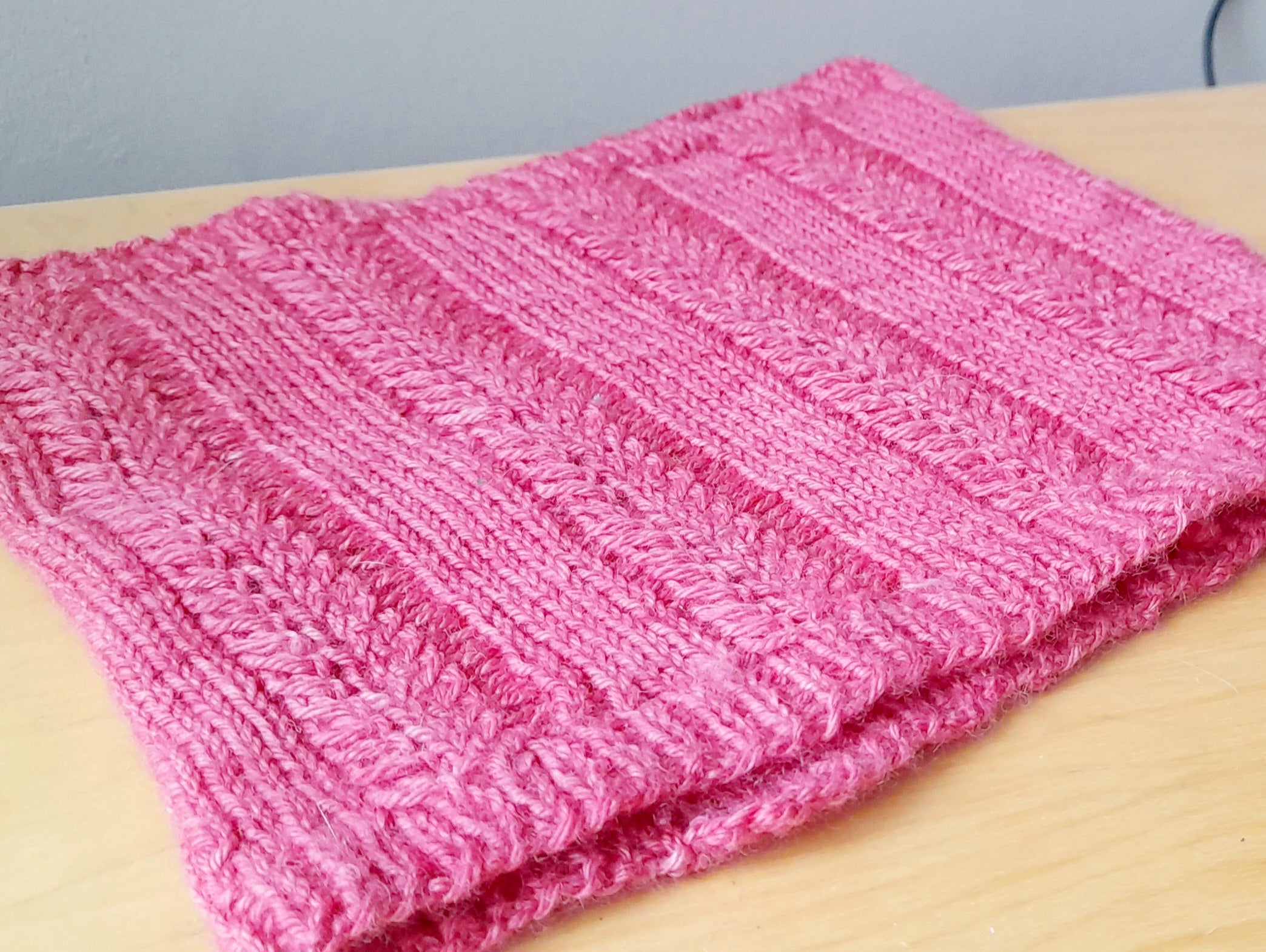 A pink cowl lying flat on a table. The cowl features blocks of textured stitches