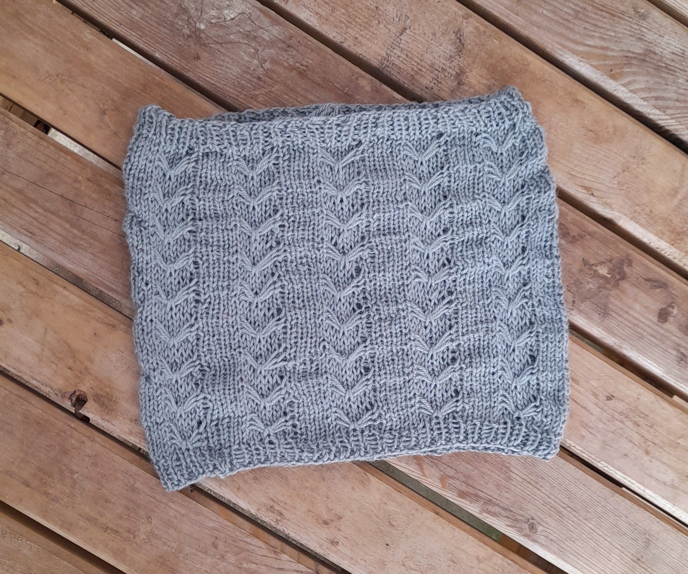 A teal cowl resting on a wooden bench. The cowl features slipped stitch v shaped details throughout