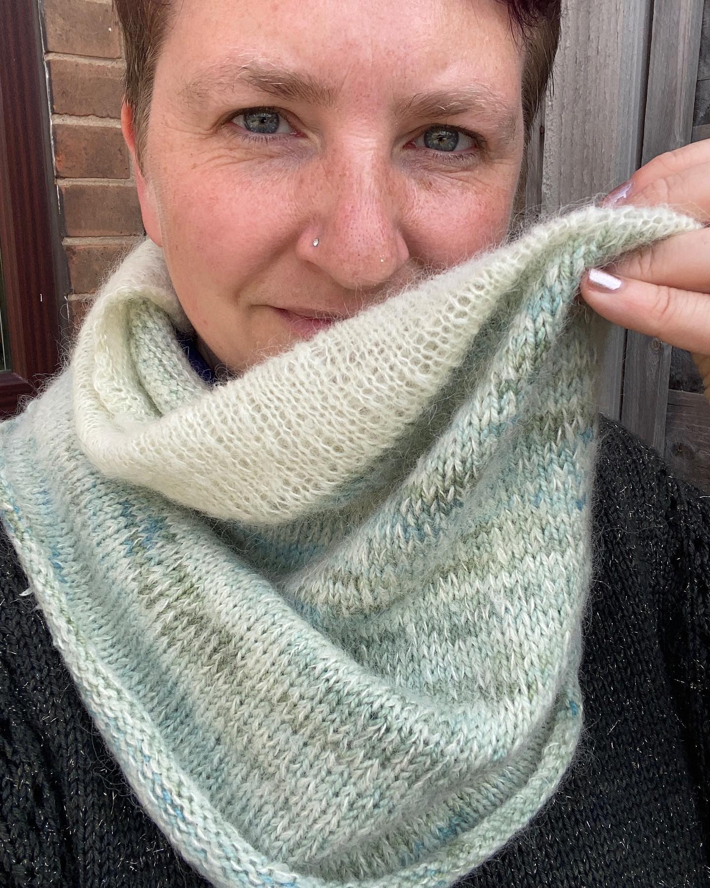 Victoria wearing a green cowl and holding up one side of it to show the variegated colour of the yarn