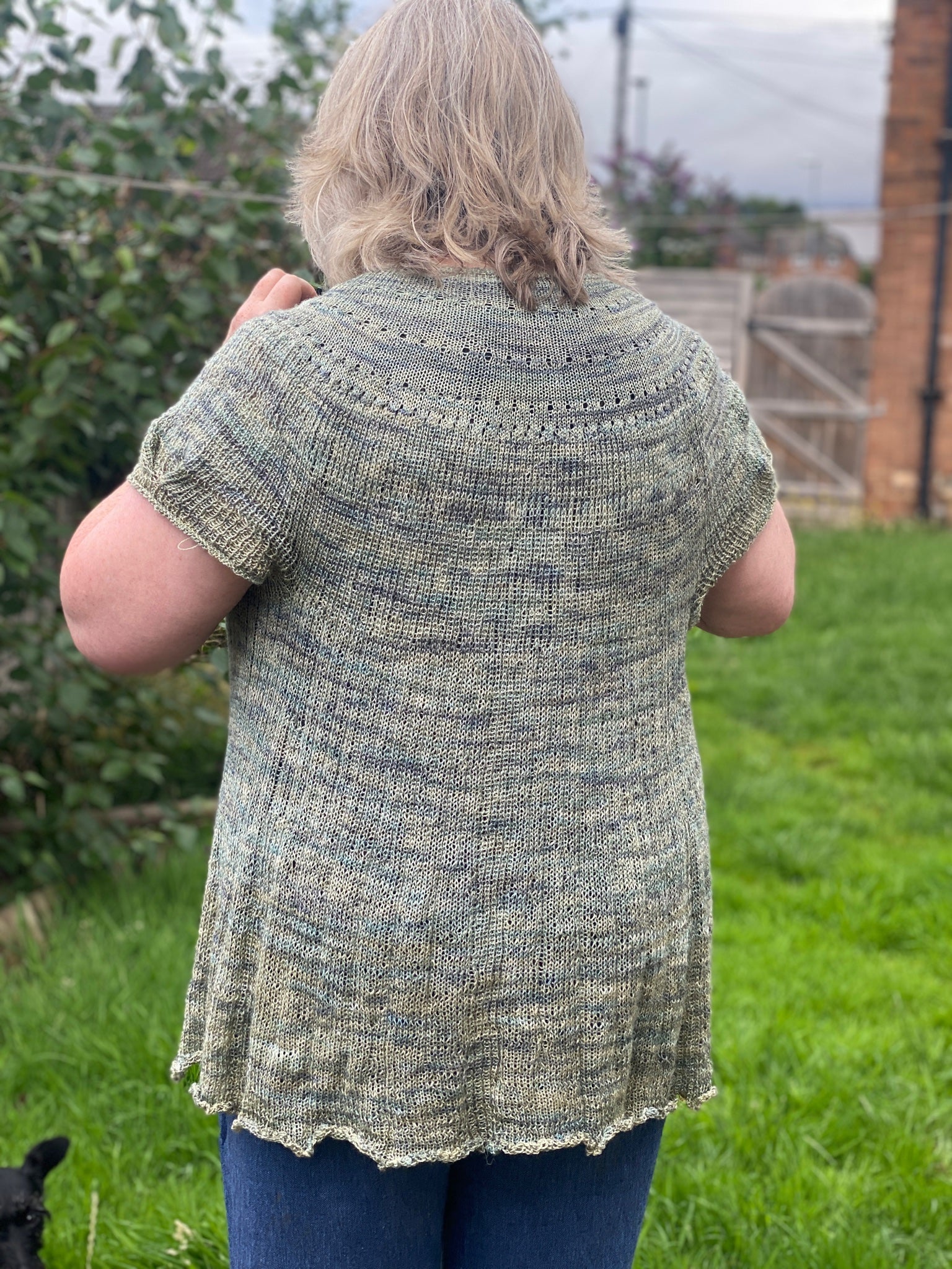 Claire facing away from the camera in a garden. She is wearing a knitted short sleeved cardigan in muted shades of green and blue. The yoke features lace detailing and the sleeves include a dart detail