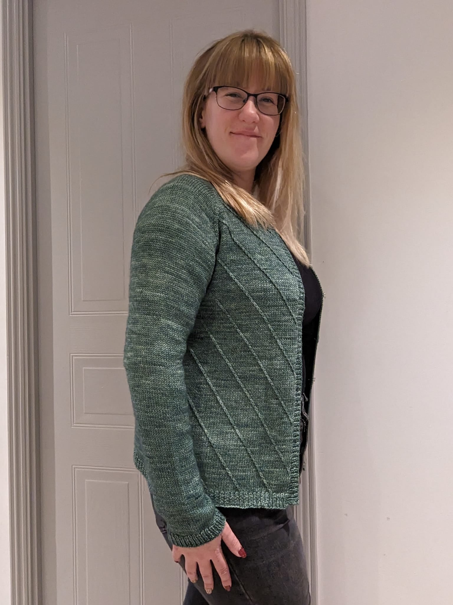 Jo standing sideways on to the camera wearing a green knitted cardigan with diagonal textured stripes on the body