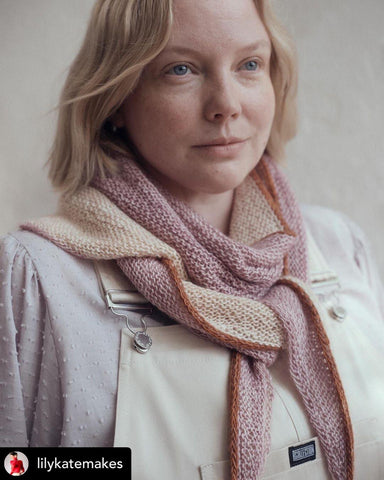 A person wearing a scarf tied like a neckerchief around their neck. The shawl is pale orange, pale purple and features an orange edging