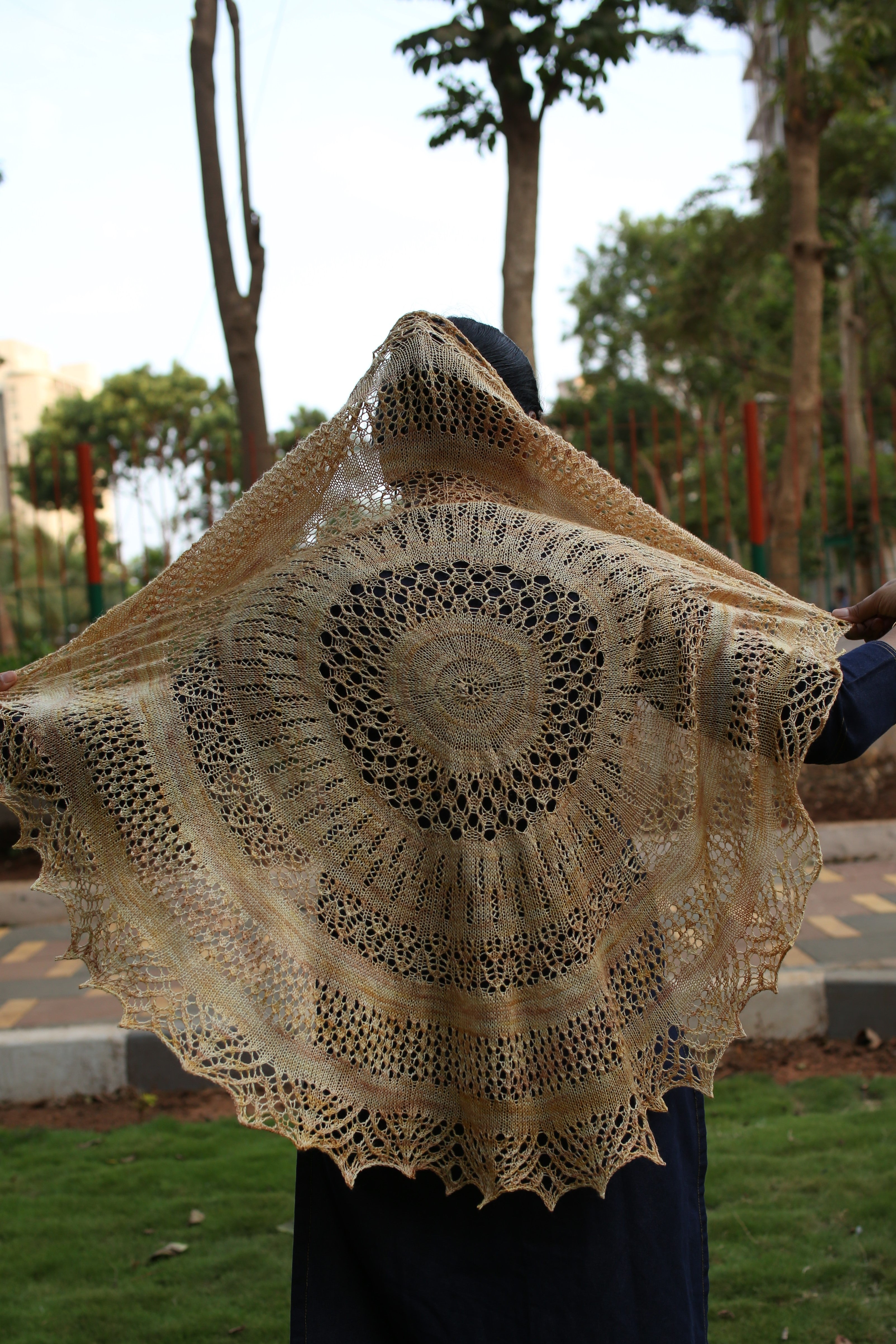 A person facing away from the camera holding a large circular shawl. The golden yellow shawl has an intricate circular lace pattern expanding out from the centre