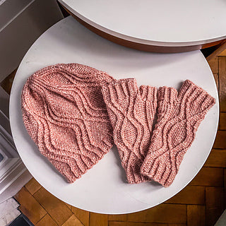 A pink cabled detailed knitted hat and mitts on a white surface