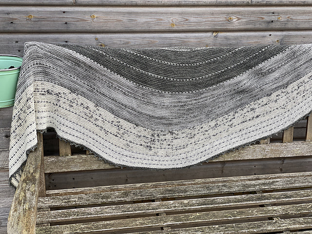 An elongated crescent shawl in shades of cream and grey arranged in a gradient from darkest at the top to lightest at the bottom