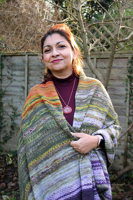 Suraya stood looking at the camera. She is wearing a wrap draped over her shoulders and is clasping the two sides together in front of her. The wrap is striped and features a wide range of colours