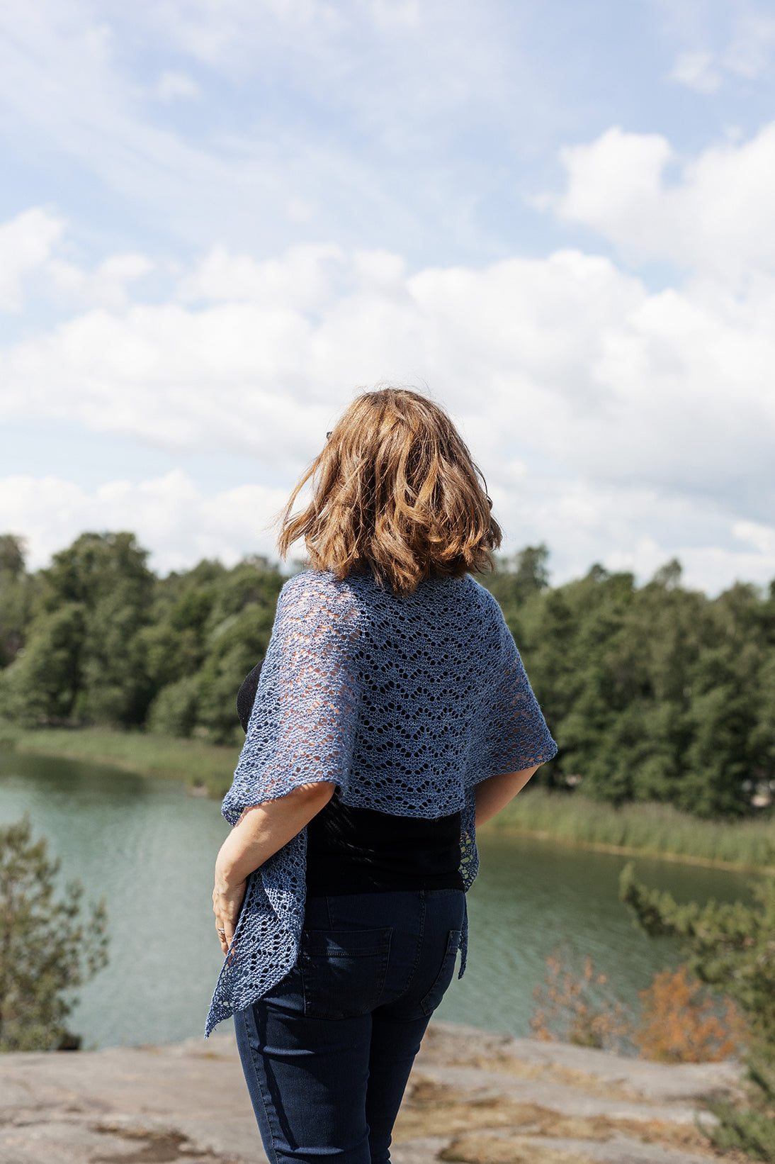 A person stands in front of a landscape including a lake and trees. The person is wearing dark jeans, a dark top and a blue knitted shawl draped across their shoulders