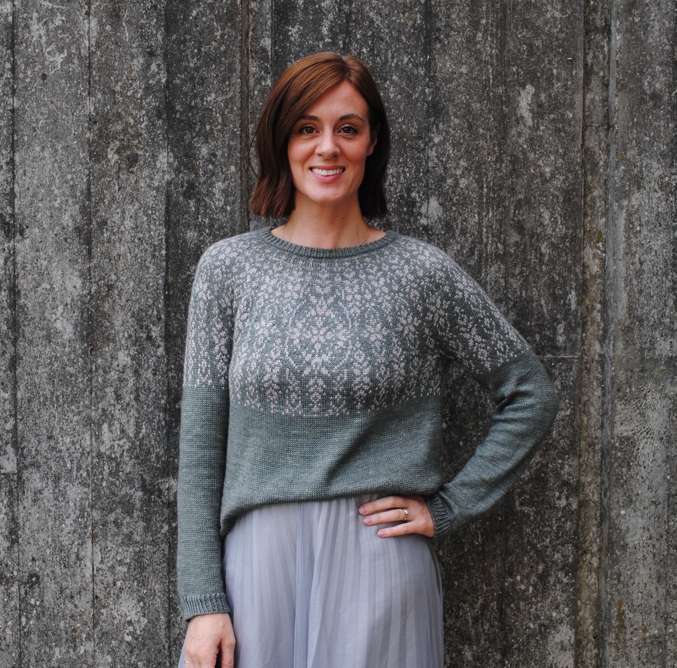 Rachel stood in front of a grey background wearing a grey skirt and a grey and beige colourwork jumper